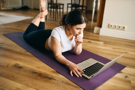 Indoor image of beautiful young woman with fit muscular body lying on her stomach on yoga mat surfing internet via laptop after home training. Barefooted girl watching online fitness video